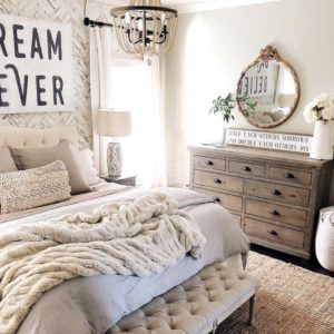Farmhouse white bedroom with cozy white and gray bedding. A bead chandelier and wood dresser. Large sign above bed that says dream believer.