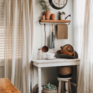 Wood shelf with flower pots and watering pale. Old vintage clock on wall and a small distressed side table with antique scale and small white stool.